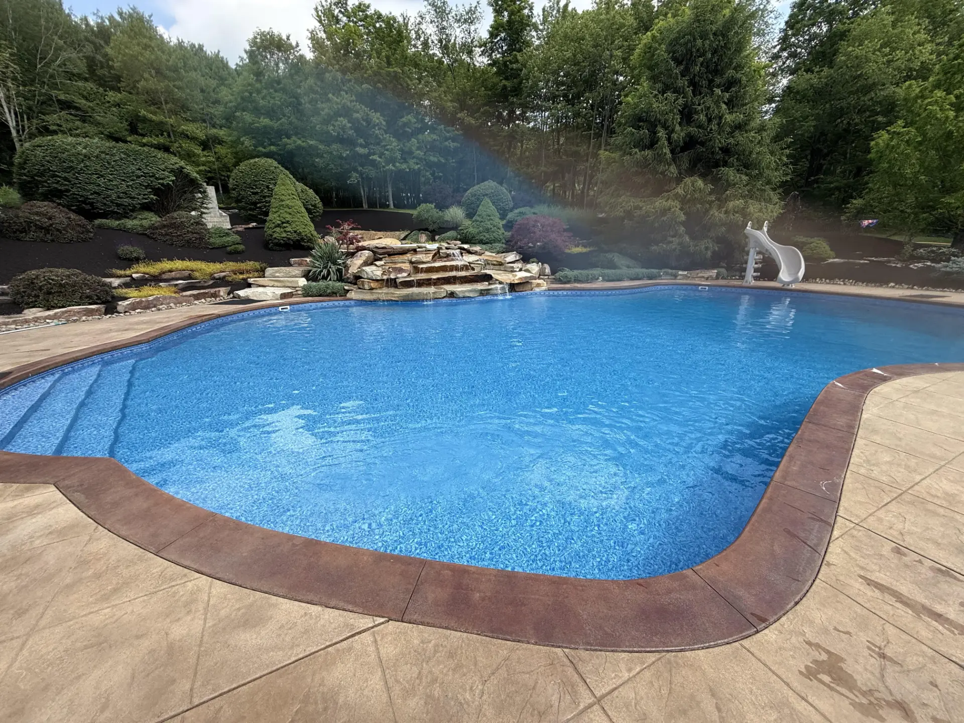 Blue backyard swimming pool with a stone waterfall and tree-lined landscape
